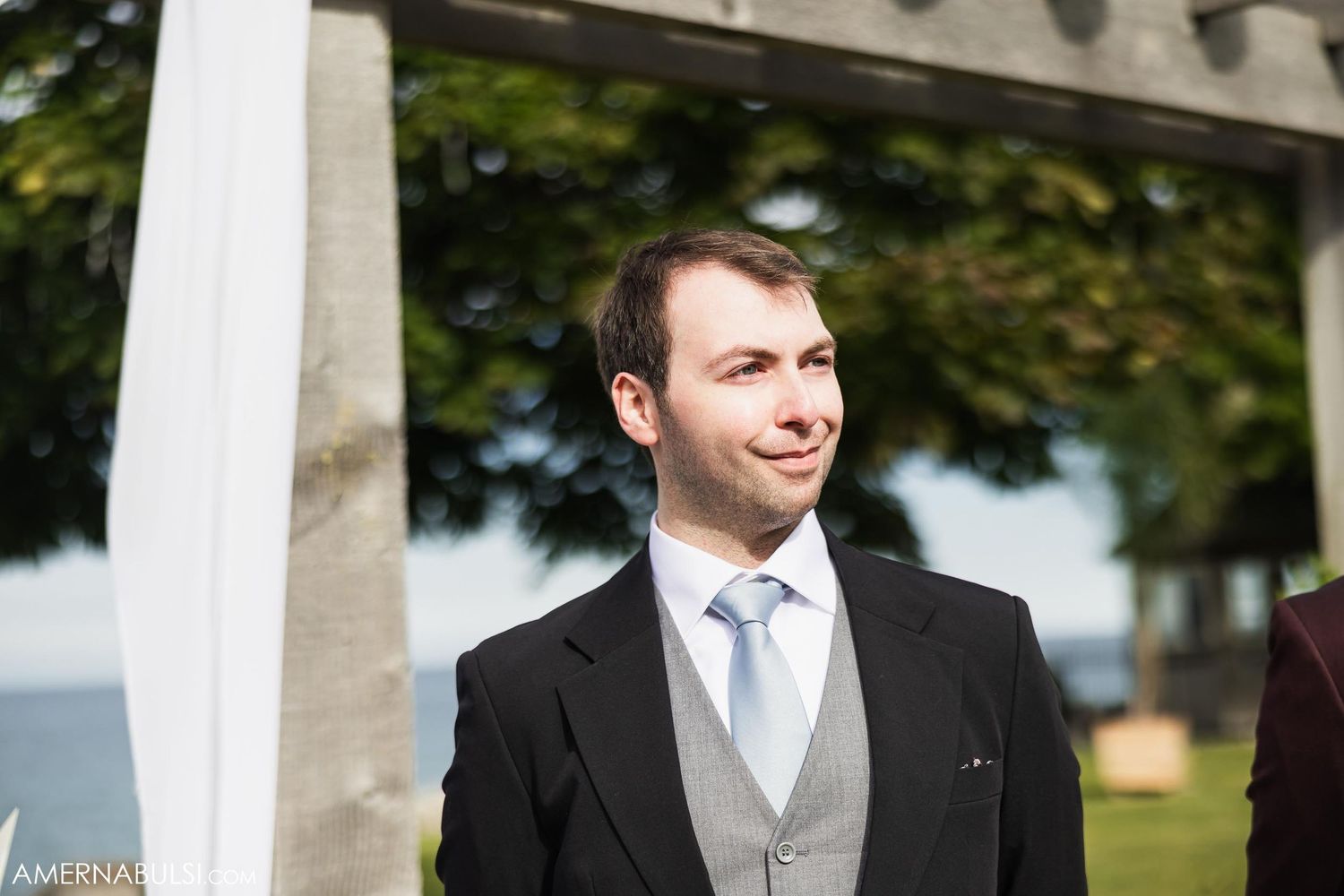 Close-up of groom in black suit and light blue tie standing under wedding pergola.