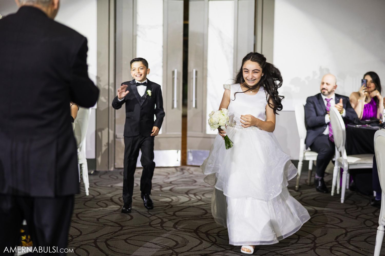 A bride walks down a carpeted hallway at her wedding while guests capture the moment.