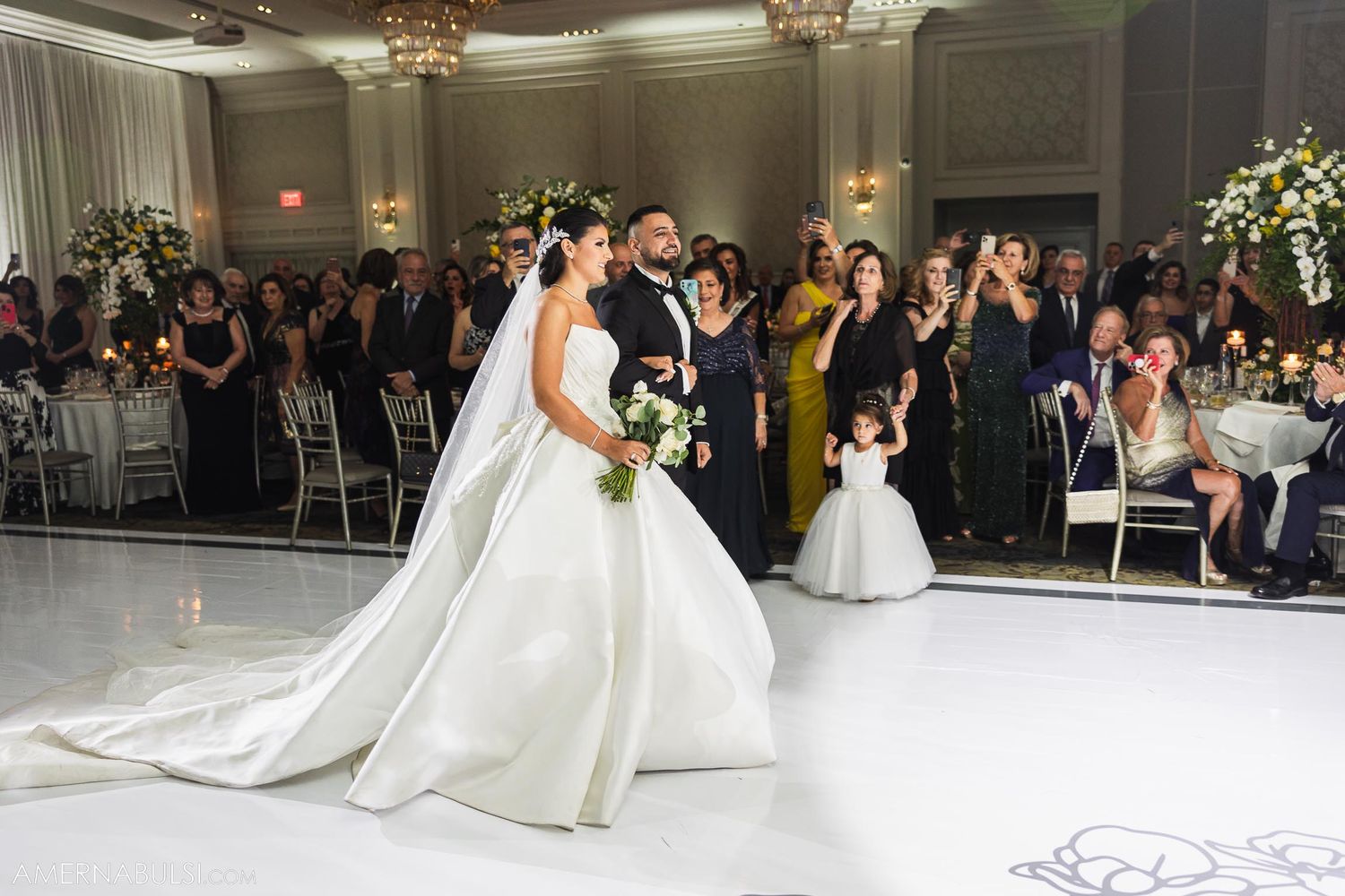 Wedding couple enters ballroom with guests cheering and flower girl following behind.