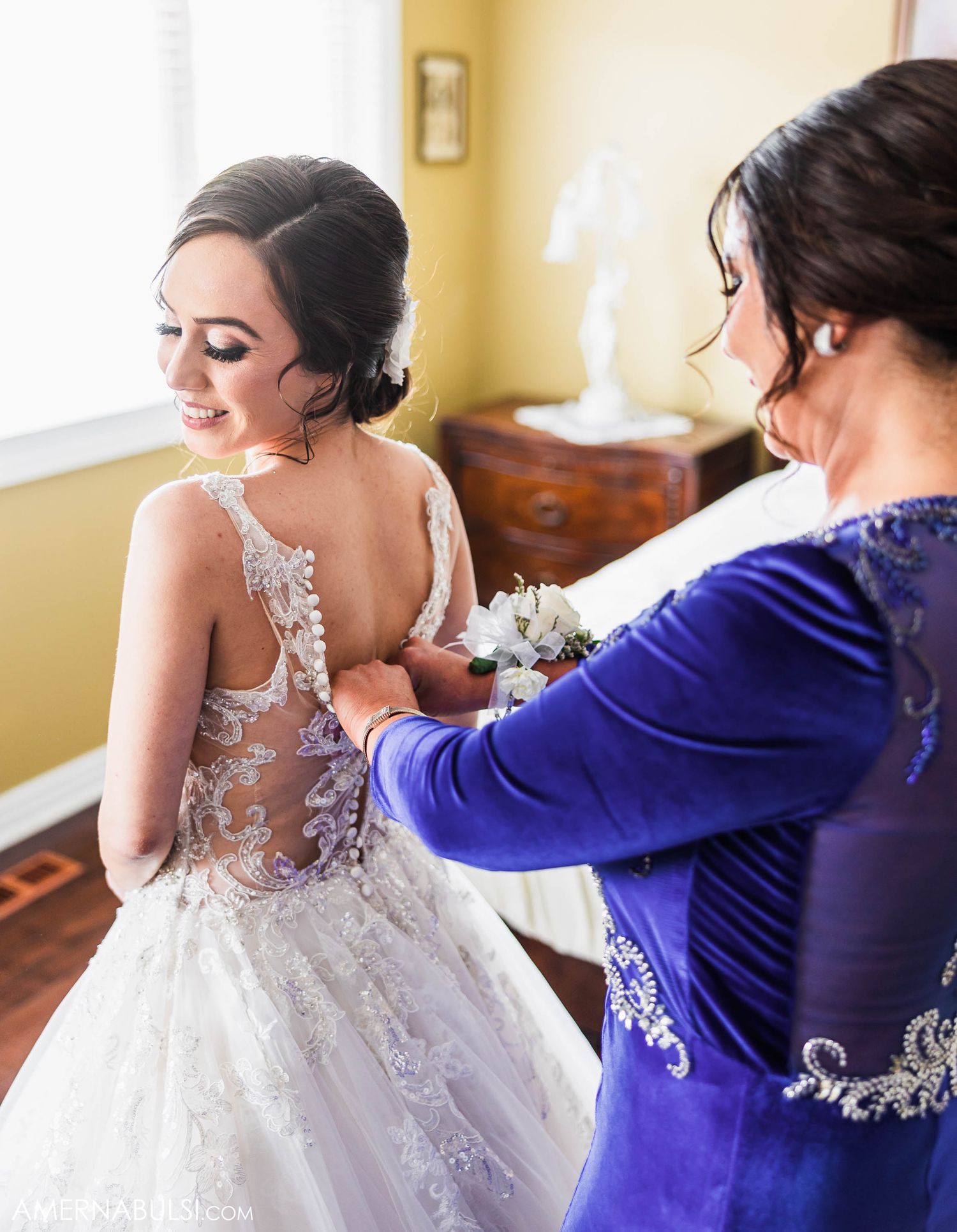 A wedding dress is being buttoned up in the back during preparation in a sunlit yellow room.