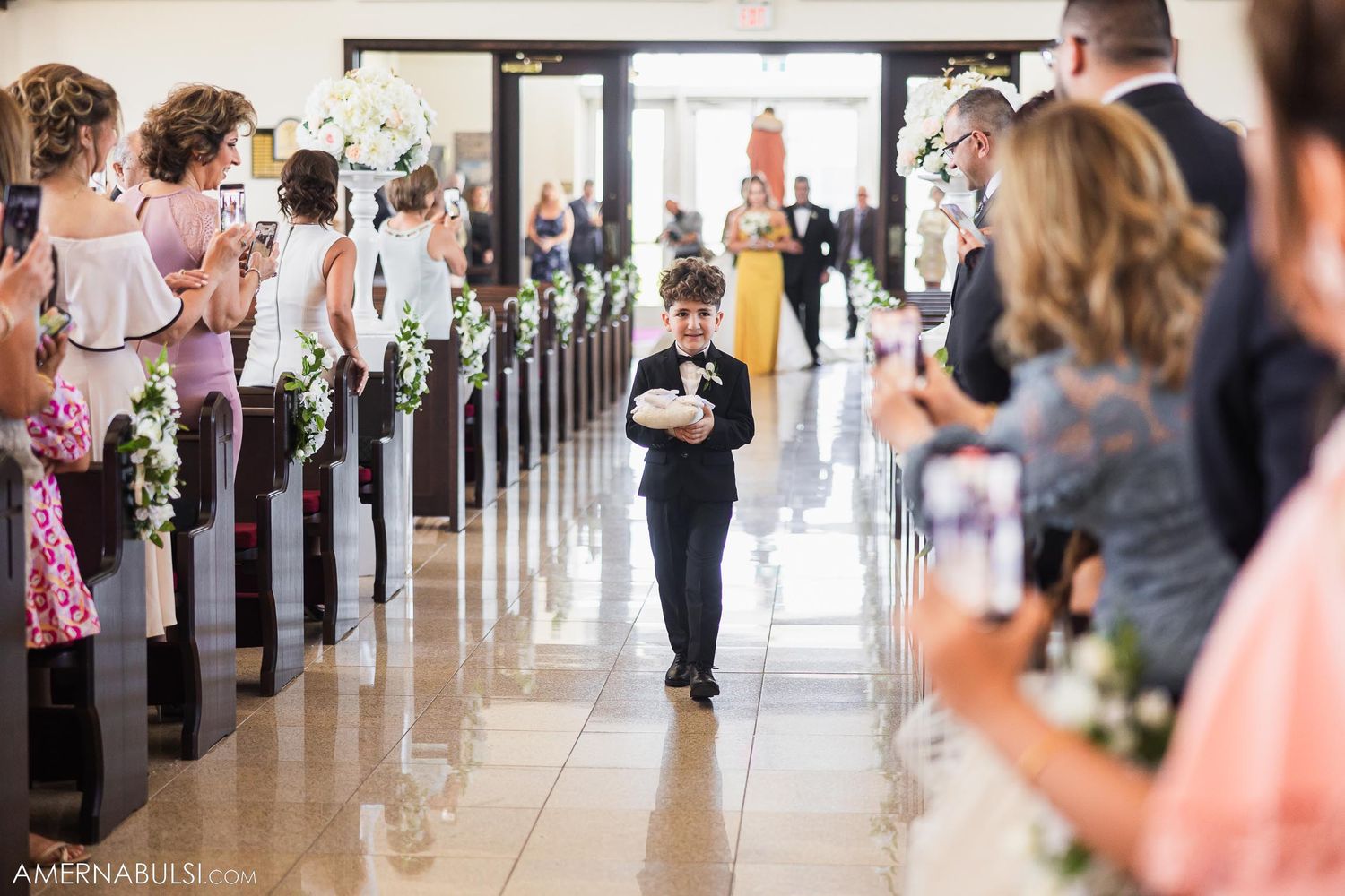 A young ring bearer walks down a church aisle decorated with floral arrangements while wedding guests look on.
