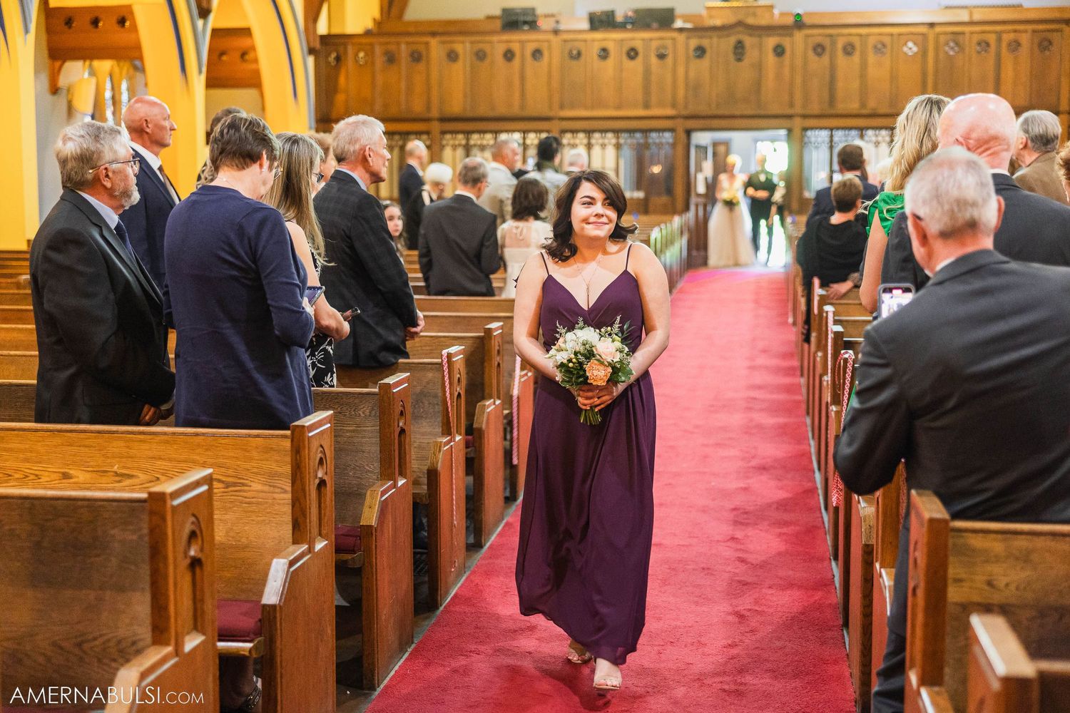 A bridesmaid in purple dress walks down church aisle with red carpet.
