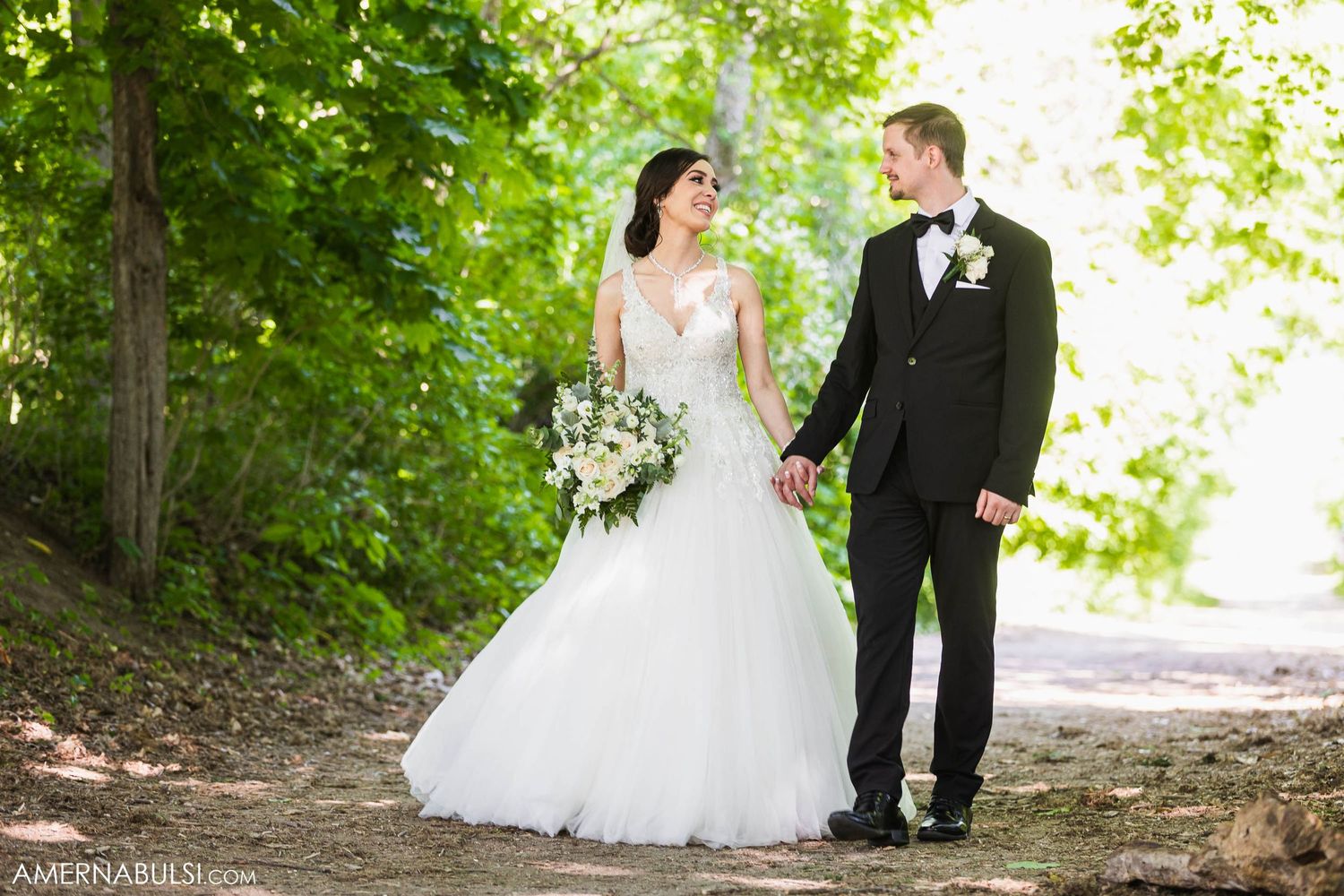 Bride and groom walking together on a sunlit woodland path with lush greenery.
