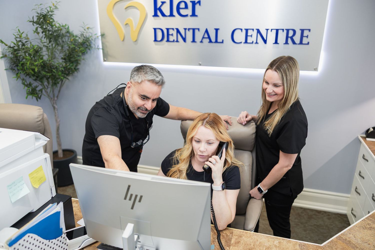 Three dental staff members collaborating at the front desk of Kler Dental Centre, reviewing information on a computer.