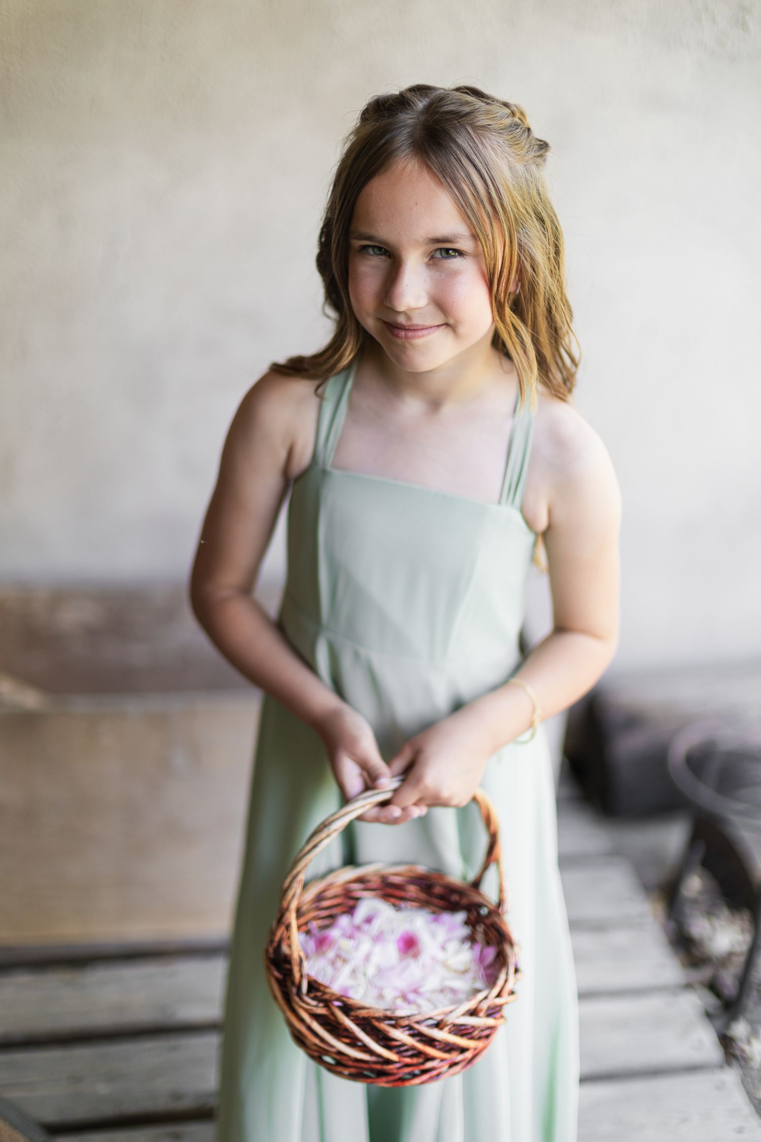 Young flower girl in sage green dress holding wicker basket with pink petals, smiling at camera.