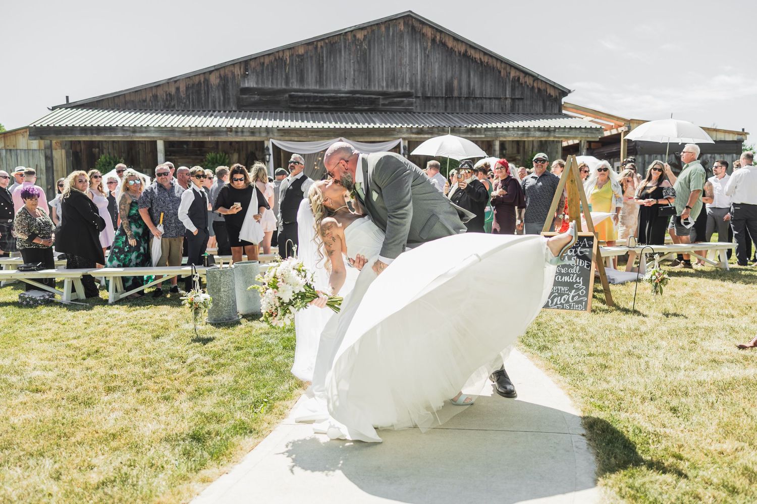 Groom dips bride in white gown during outdoor wedding ceremony with guests watching near rustic barn.