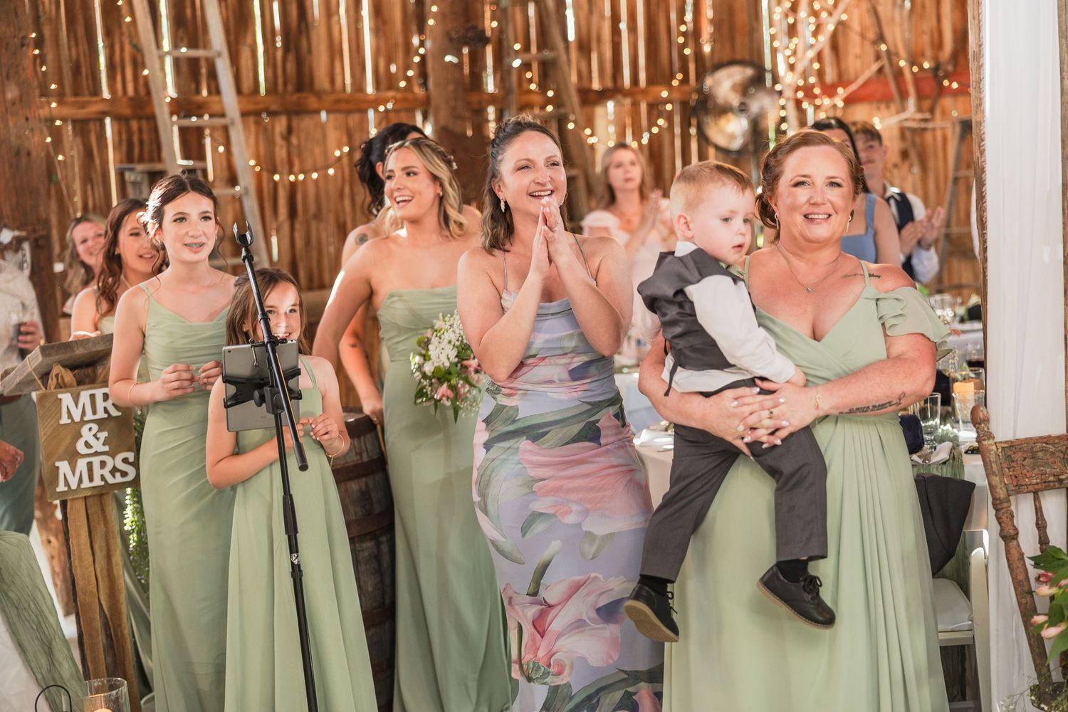 Bridesmaids in sage green gowns react joyfully at a rustic barn wedding with Mr & Mrs sign.