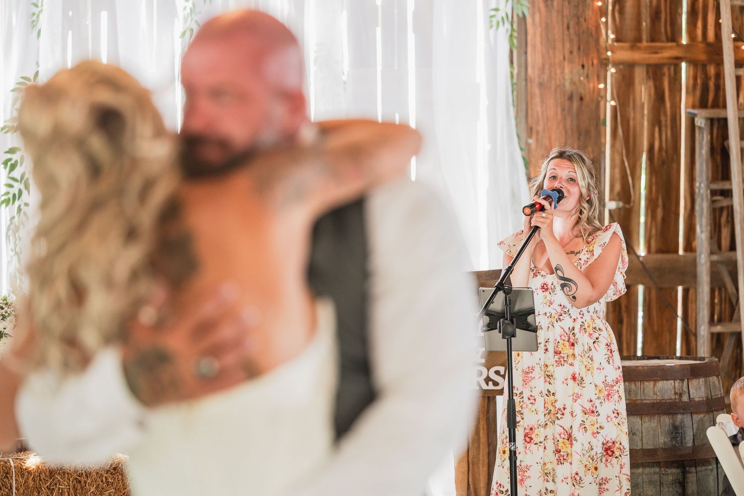 Woman singing into microphone at rustic barn wedding reception while couple shares first dance.