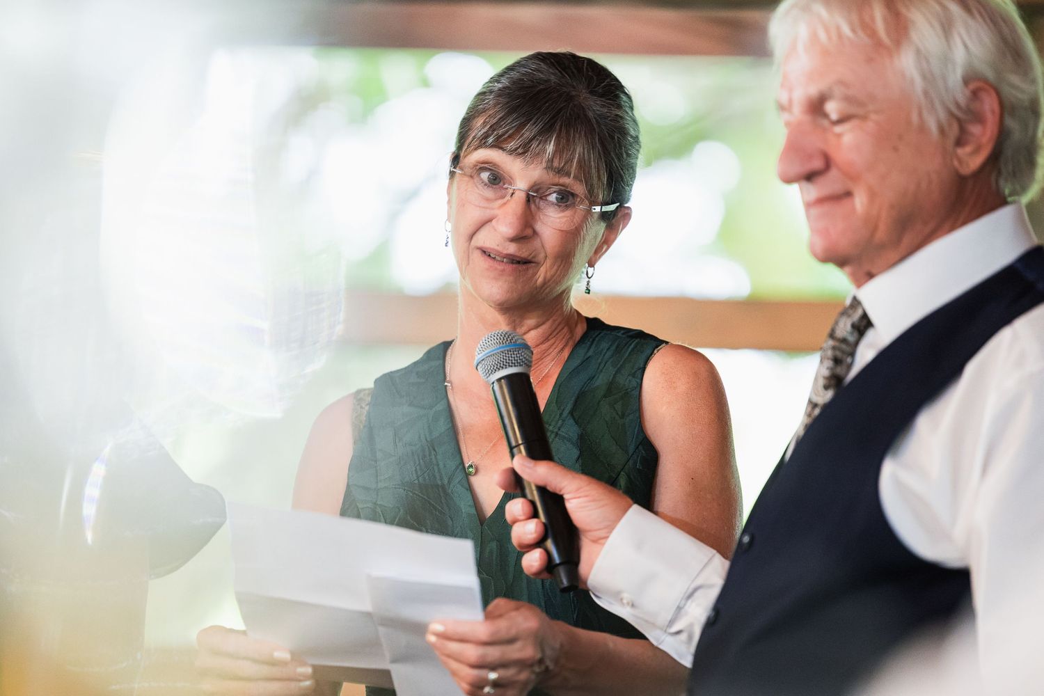 Woman in green dress speaks into microphone at wedding reception while man in suit holds papers beside her.