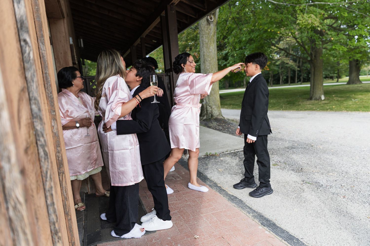 Bridesmaids in pink robes gather under a wooden shelter while a groomsman in a black suit gestures outdoors.
