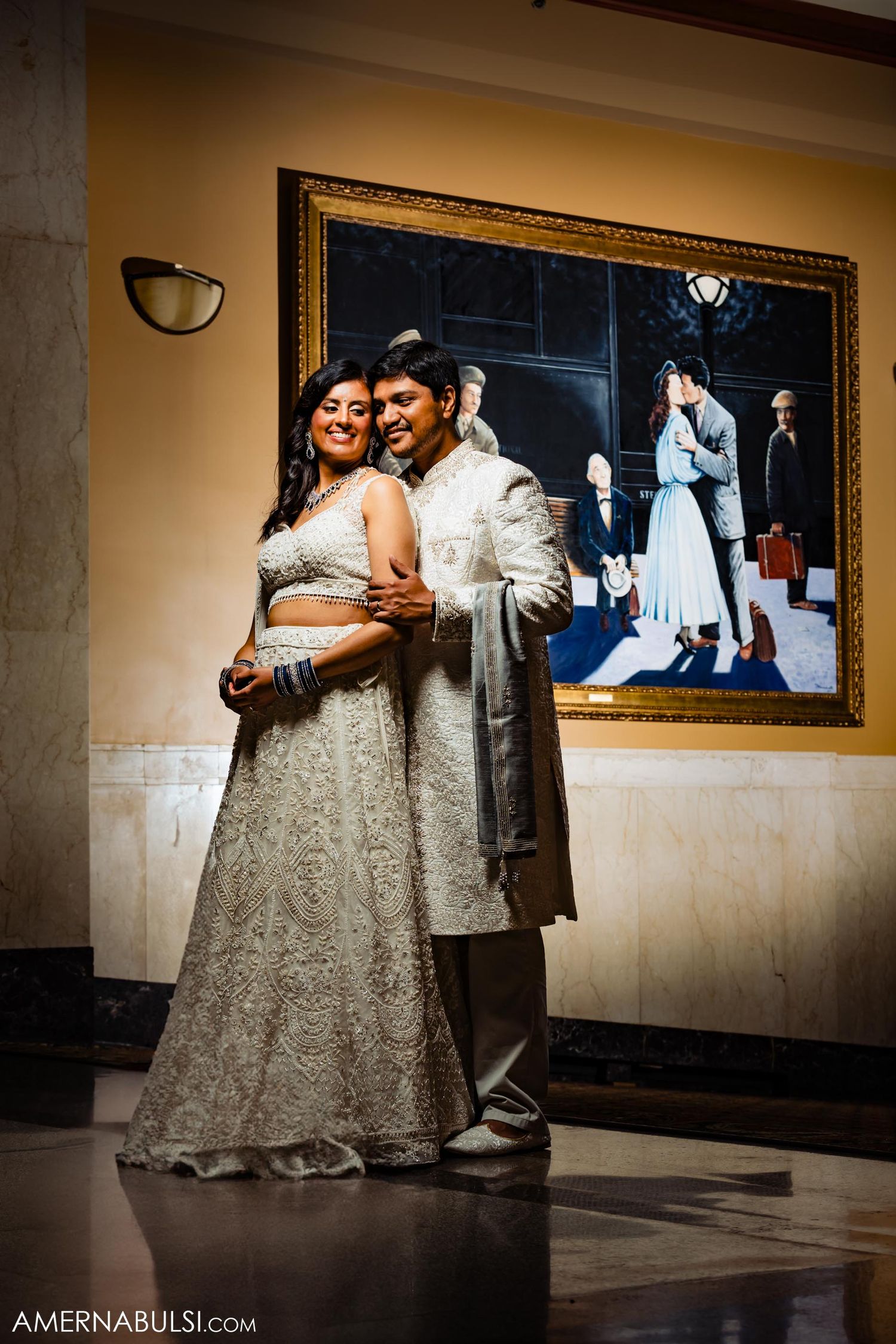 A couple poses in traditional wedding attire with an ornately framed artwork in the background.