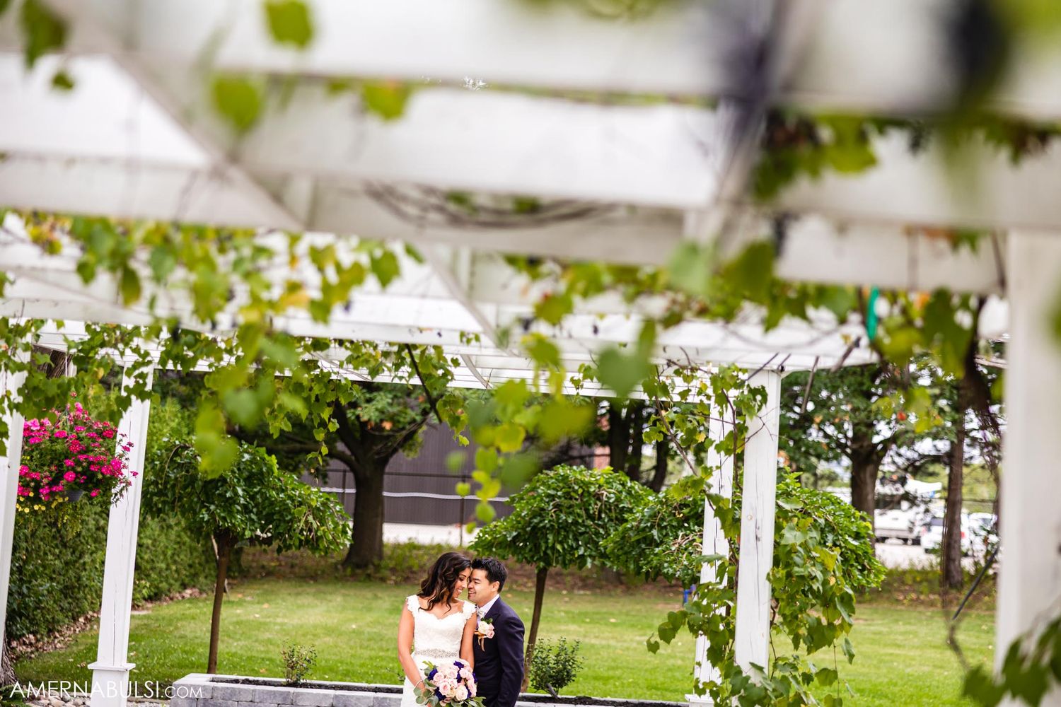 Wedding couple shares intimate moment under pergola decorated with greenery and white draping.