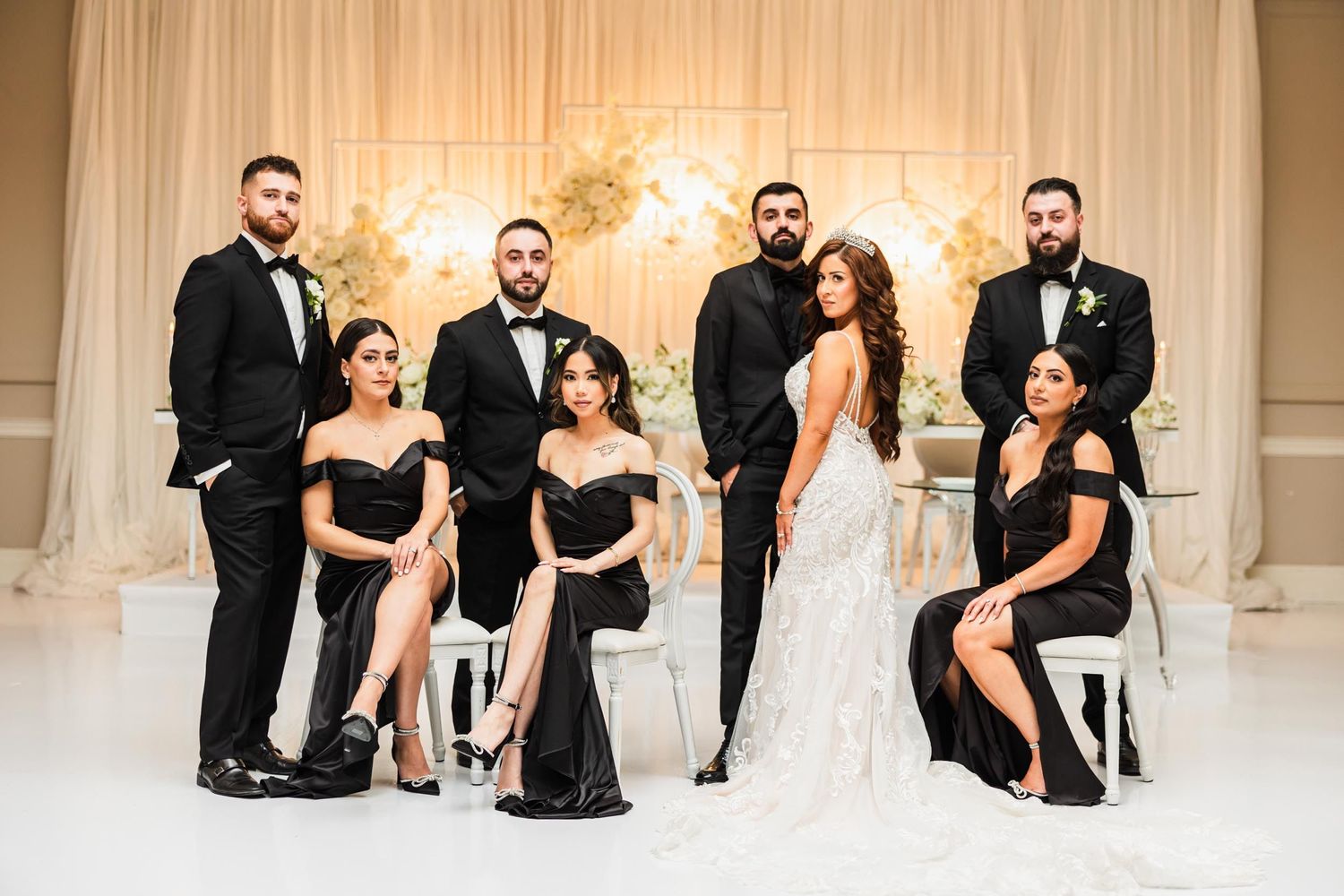 Wedding party poses in formal black attire against white backdrop with floral arrangements.