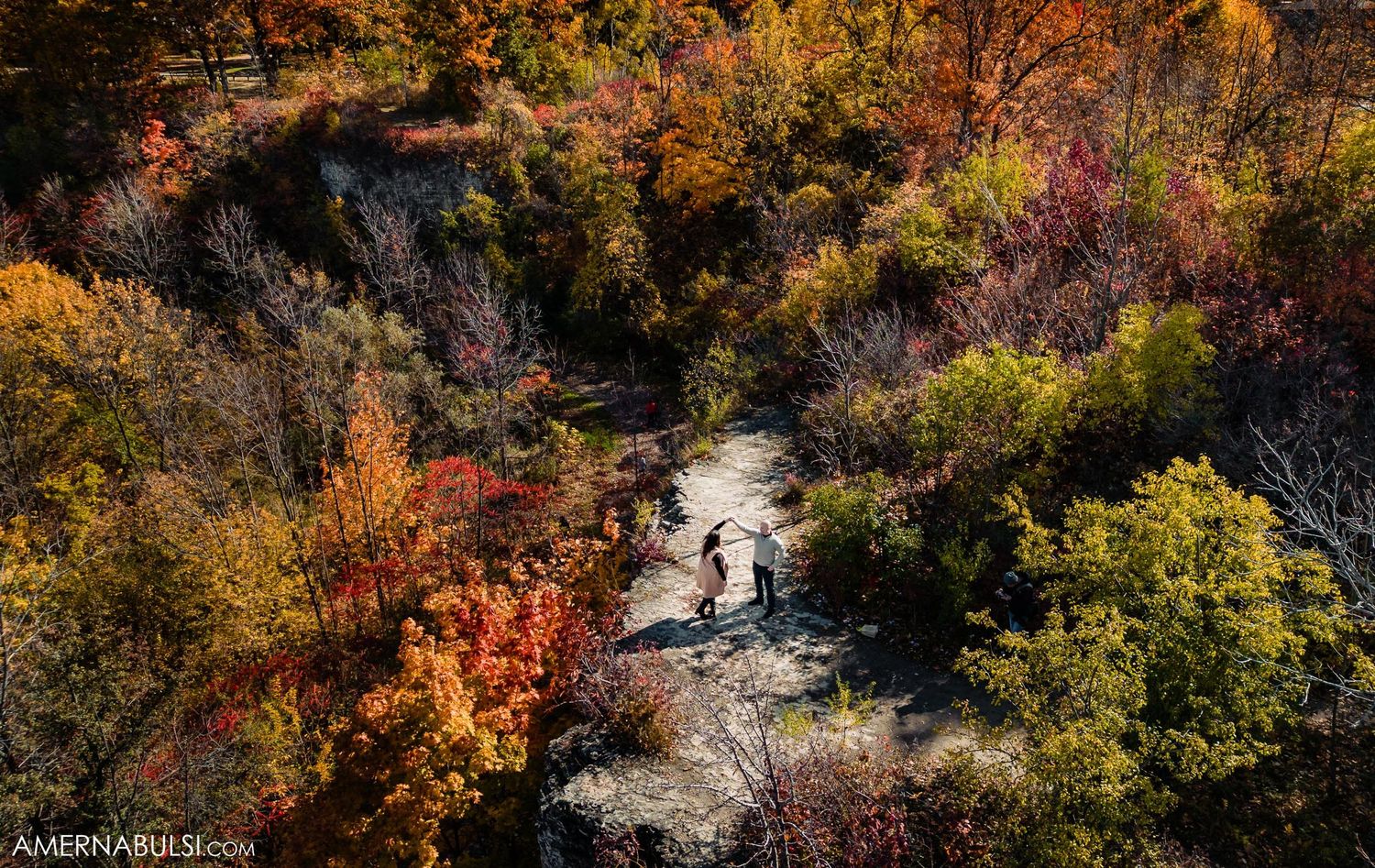 Aerial autumn view of hikers walking along a winding river trail surrounded by colorful fall foliage.