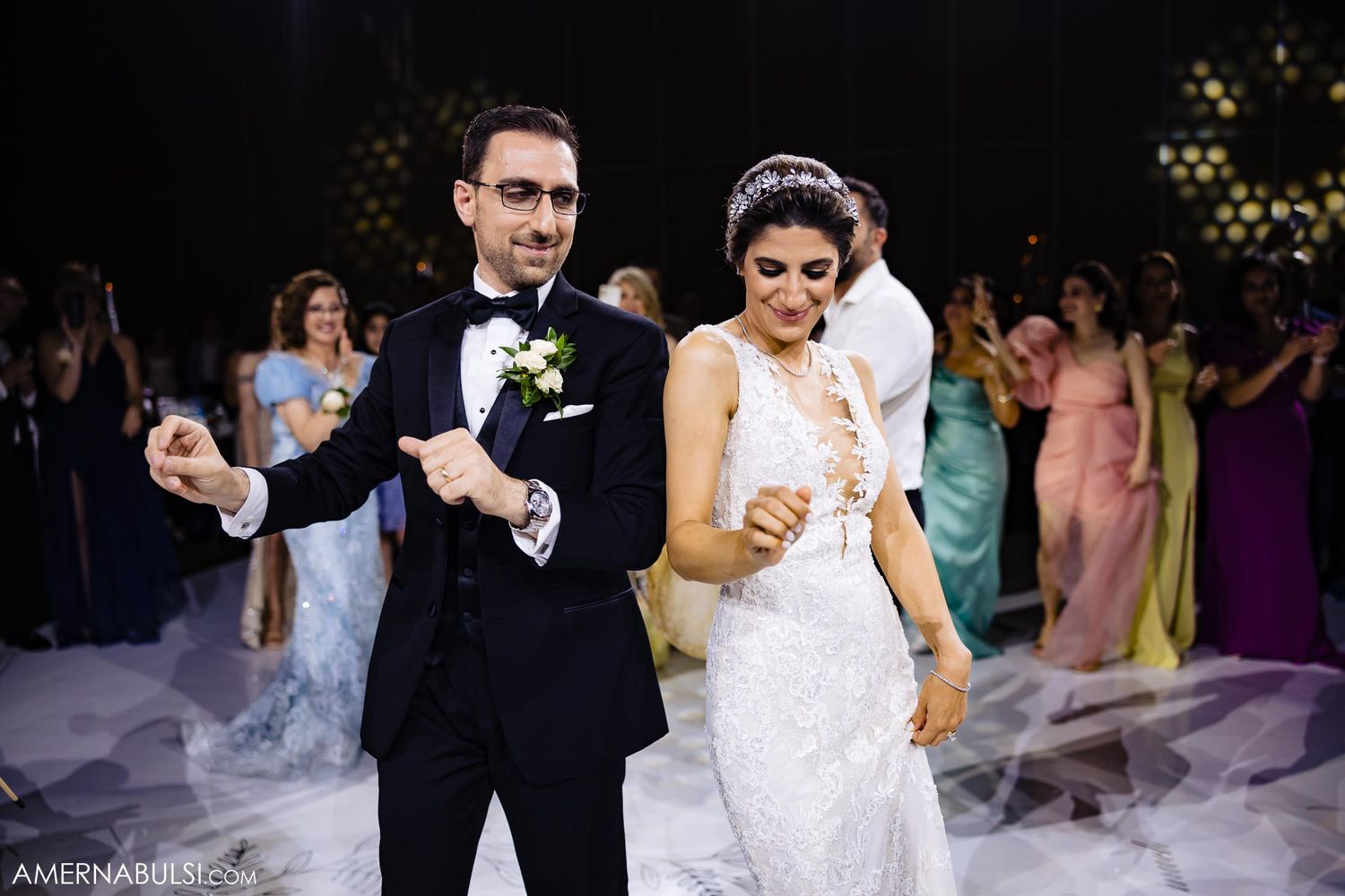 Wedding couple dances energetically surrounded by guests and colorful party lights.