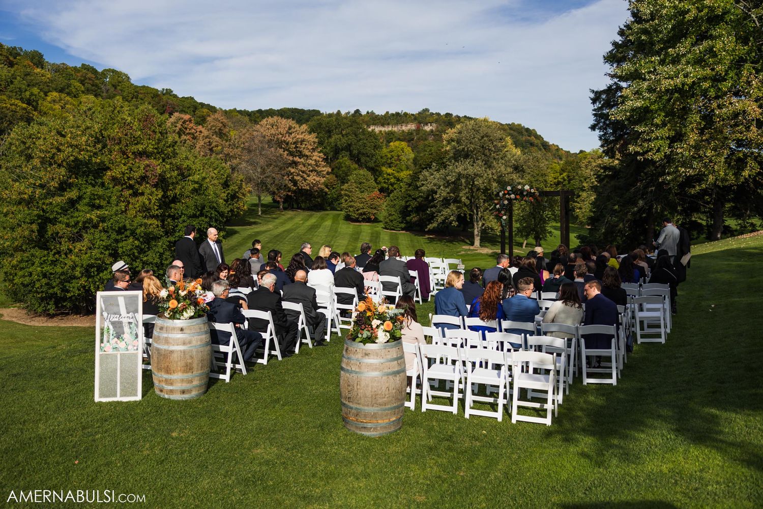 Outdoor wedding ceremony setup with white chairs and wine barrels on hillside lawn.