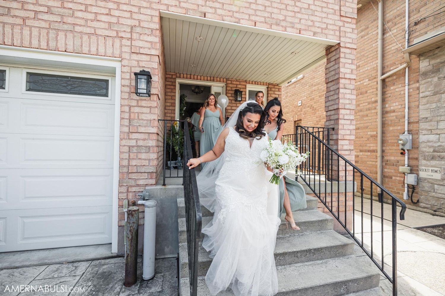 Wedding party helping with dress and luggage outside brick building entrance.