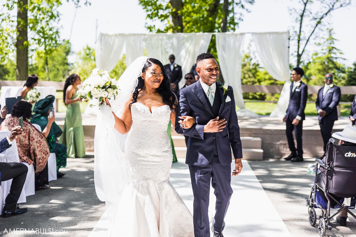 Bride and groom exit outdoor wedding ceremony under white draped arch amidst cheering guests.