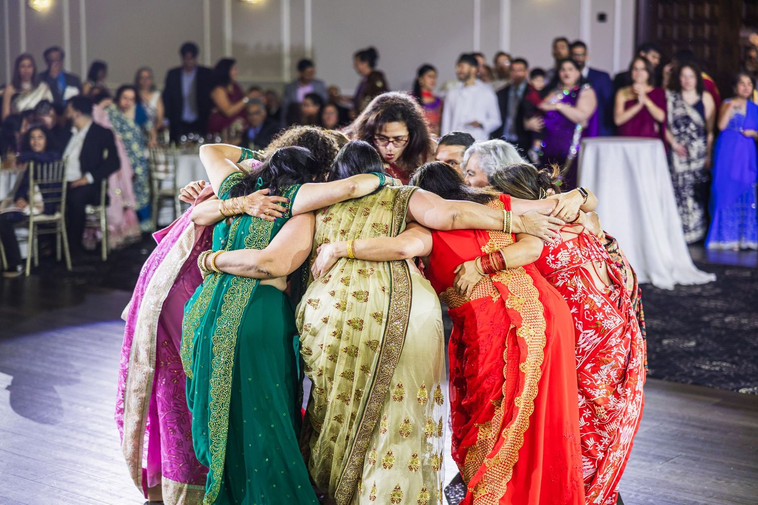 Wedding guests in colorful traditional Indian saris dance together in a line at a reception celebration.