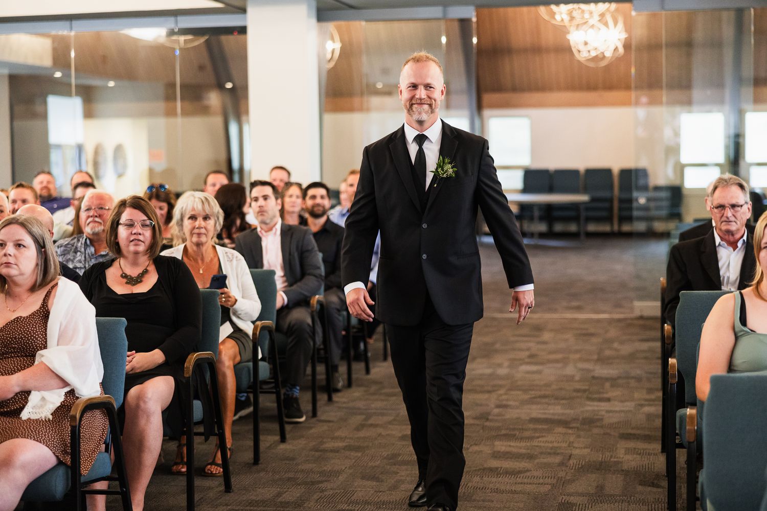 Wedding attendant walking down aisle between seated guests in formal attire.