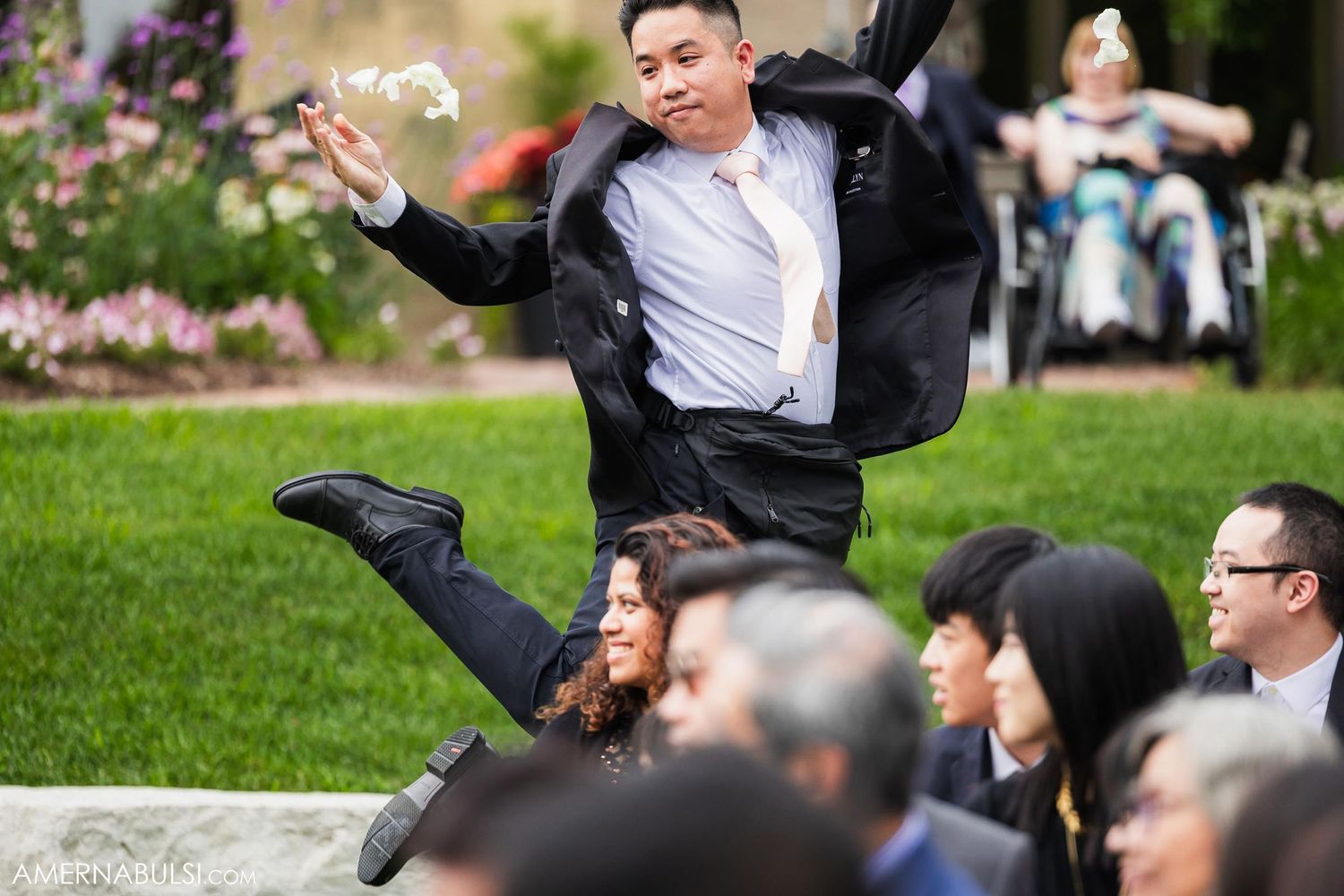 Wedding guest leaps joyfully in the air during outdoor celebration while others watch seated.