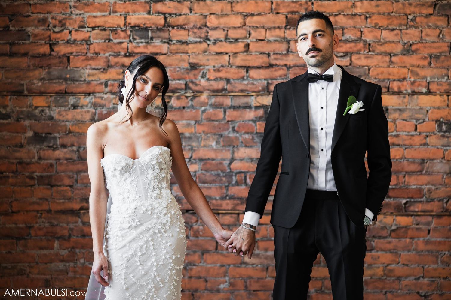 A couple stands elegantly against exposed brick in formal wedding attire.