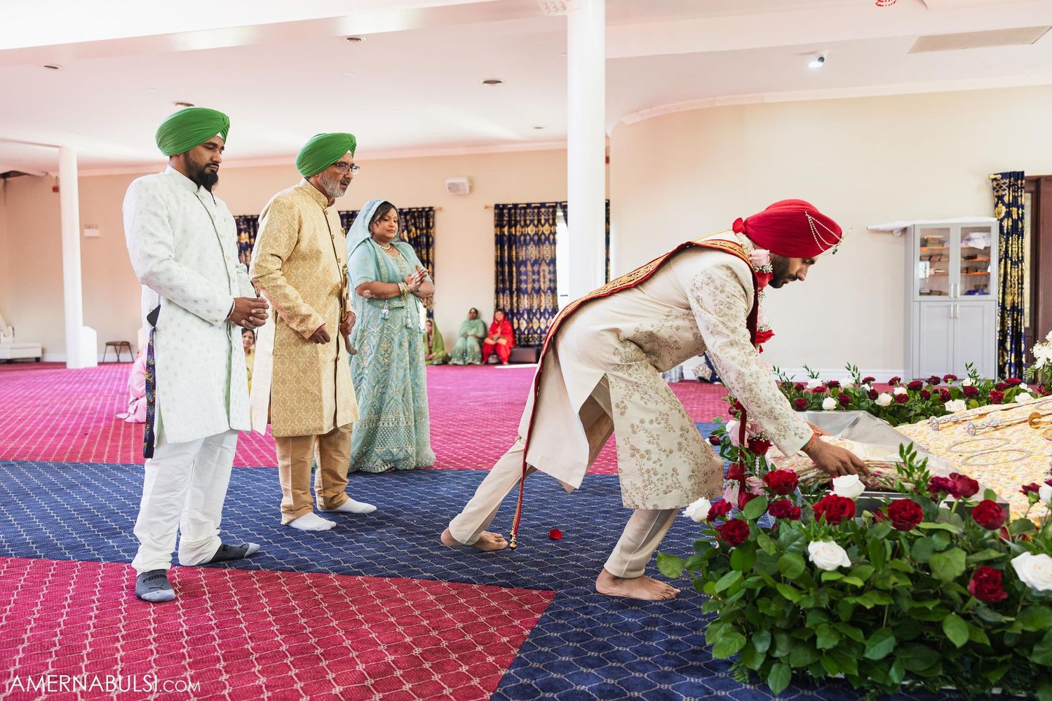 Ceremonial participants in traditional Sikh attire interacting near floral decorations on blue carpet.