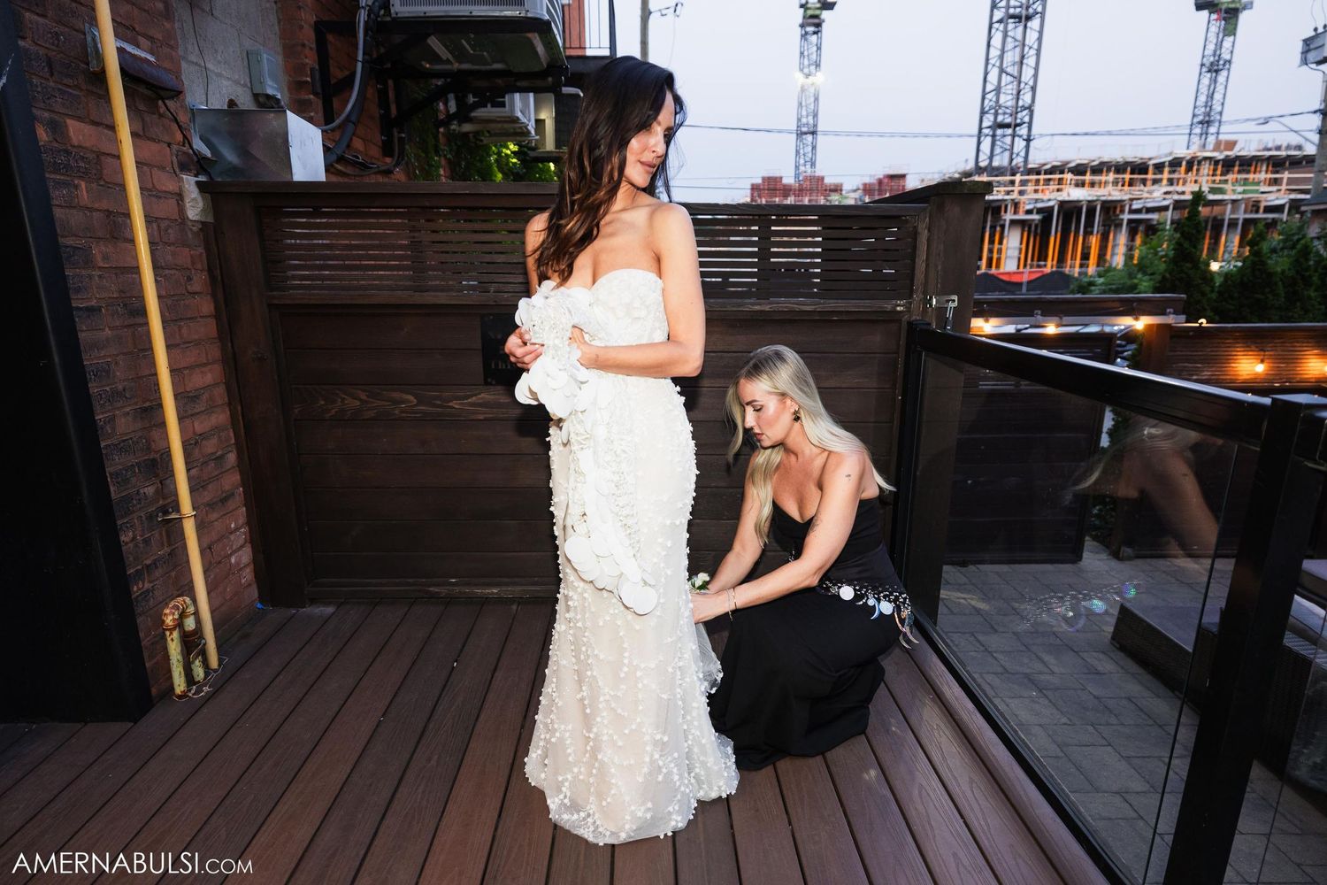 Wedding dress being adjusted on outdoor deck with industrial backdrop.