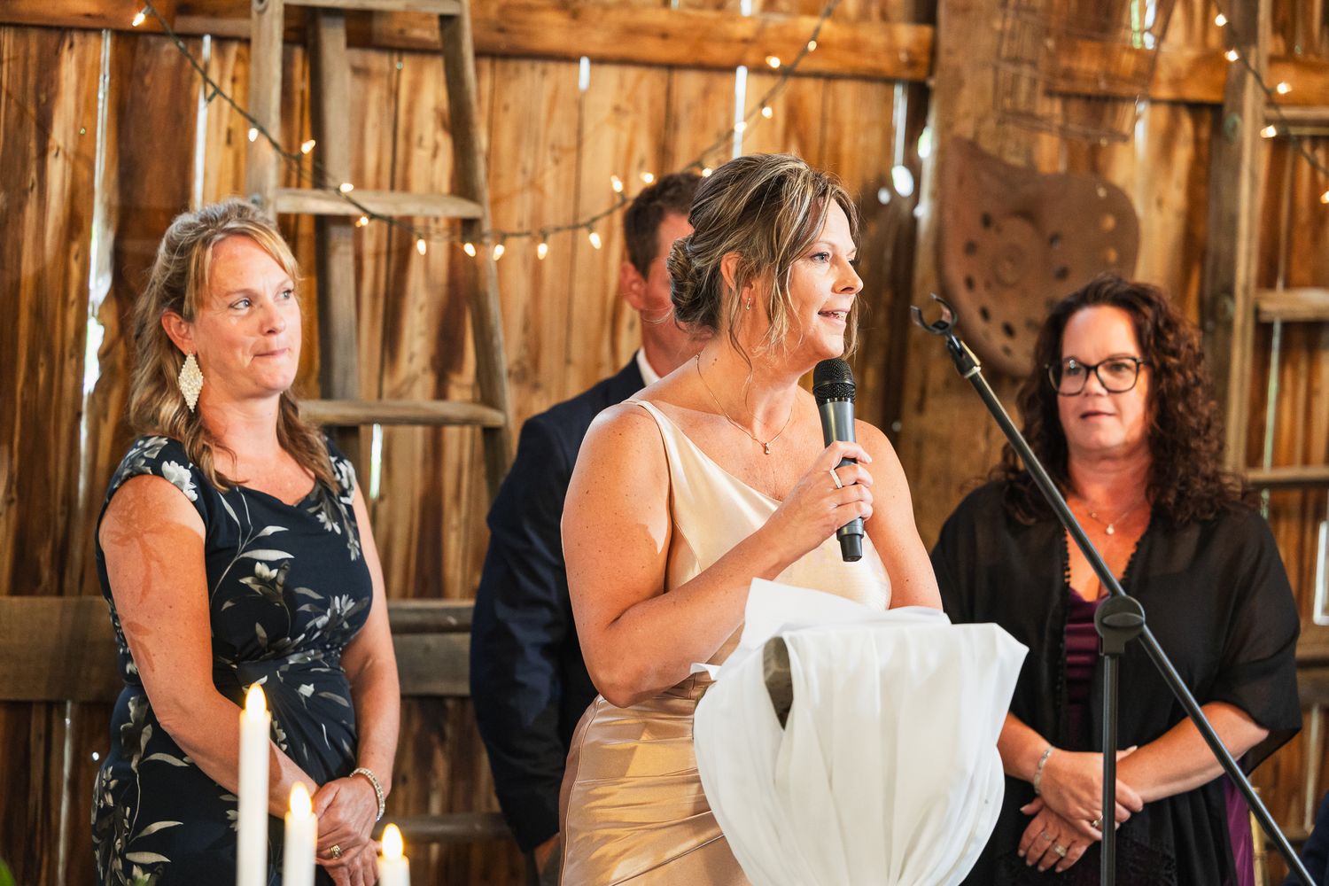 Wedding speech being delivered at rustic barn reception with string lights and wooden backdrop.