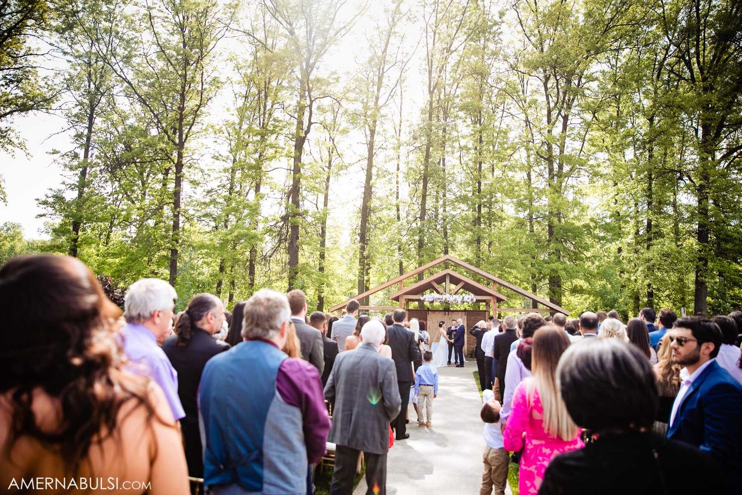Wedding guests gather outdoors under trees for a ceremony at a rustic venue.