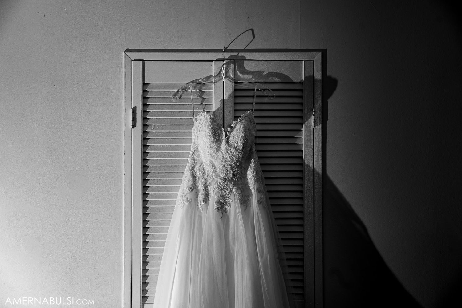 Wedding dress hanging in silhouette against white shuttered window.