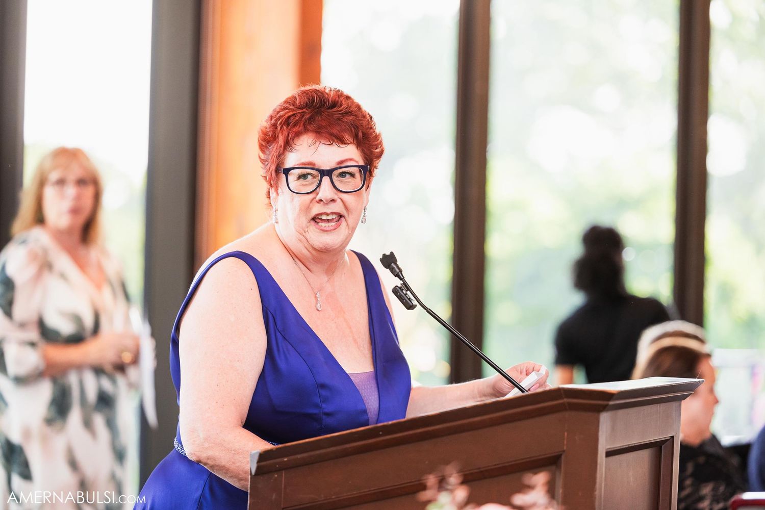Speaker in blue dress gives speech at podium during wedding reception.