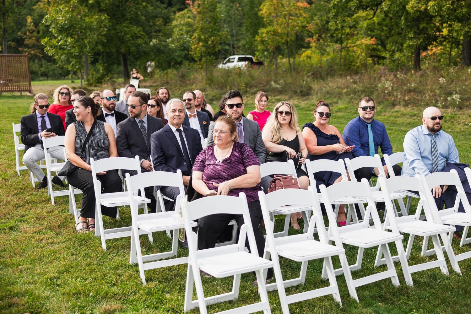 White folding chairs set up outdoors for wedding ceremony with guests seated under autumn trees.