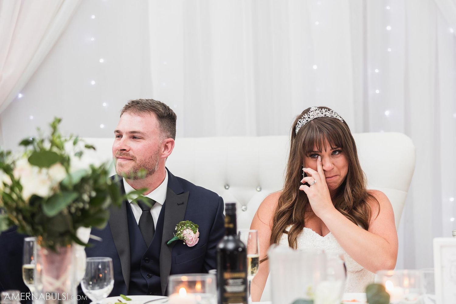 Bride shows emotional reaction during reception speech while seated next to groom.