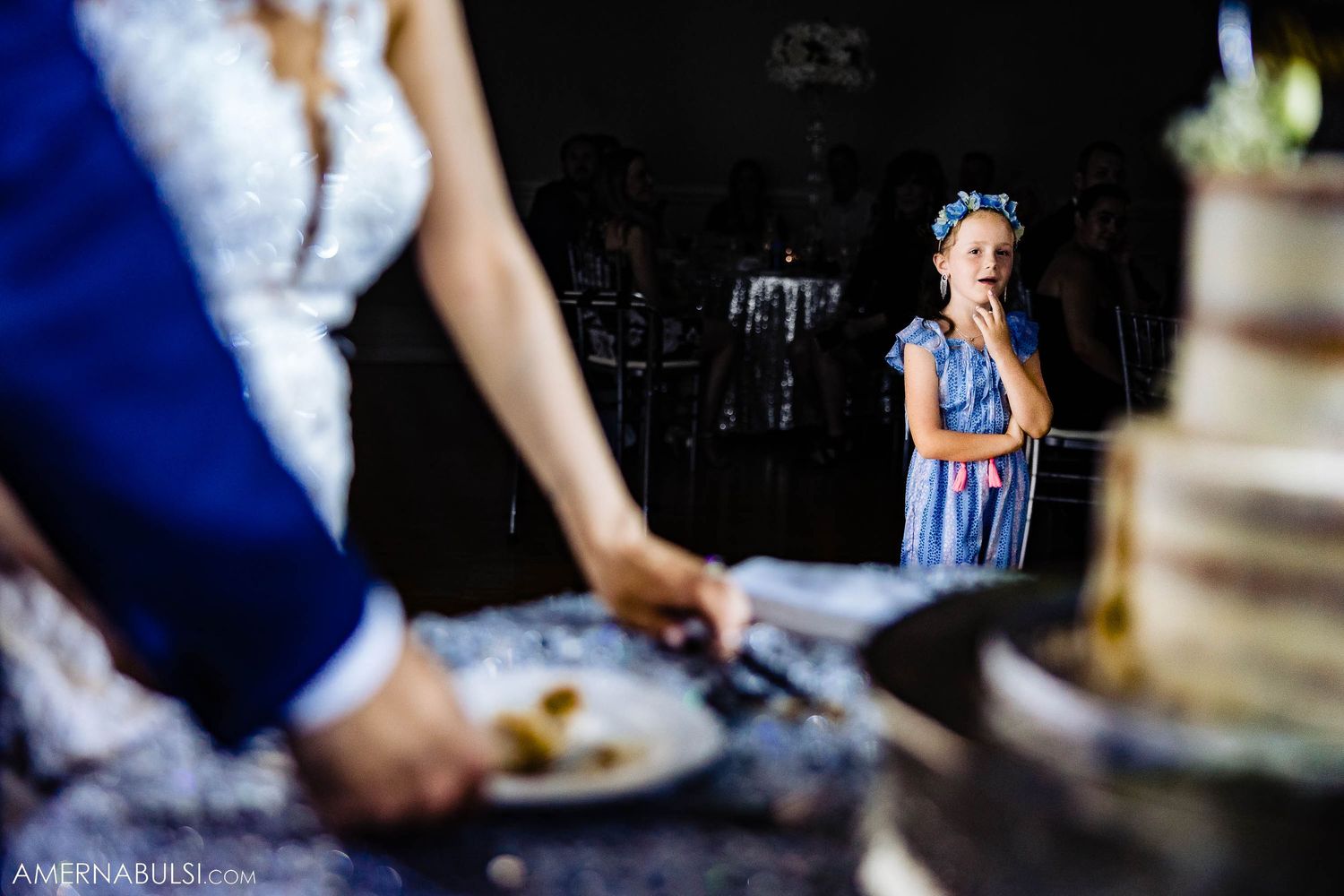 Wedding cake cutting moment while a young guest in blue dress watches from the background.