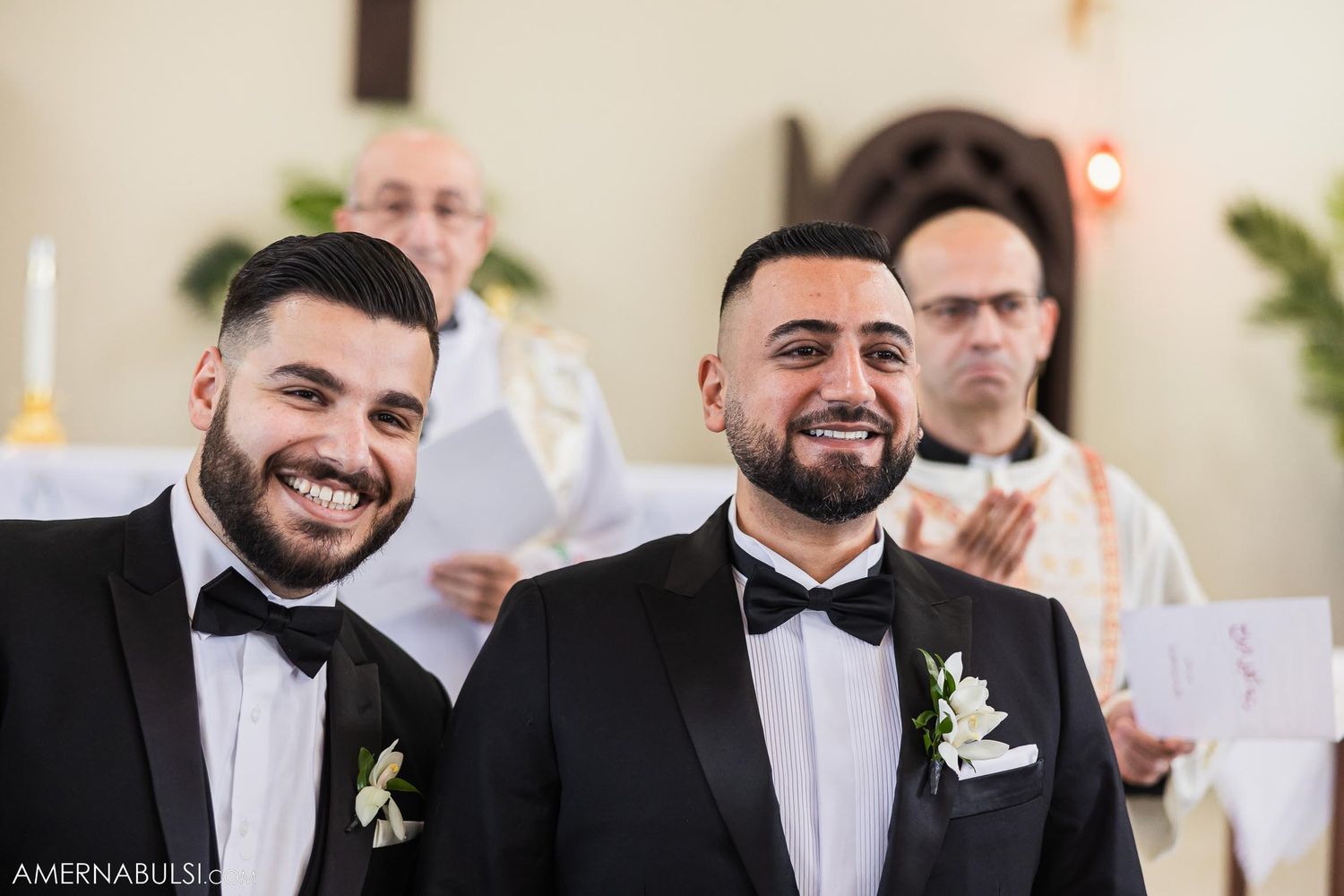 Two grooms in matching black tuxedos share happy moment at altar during ceremony.