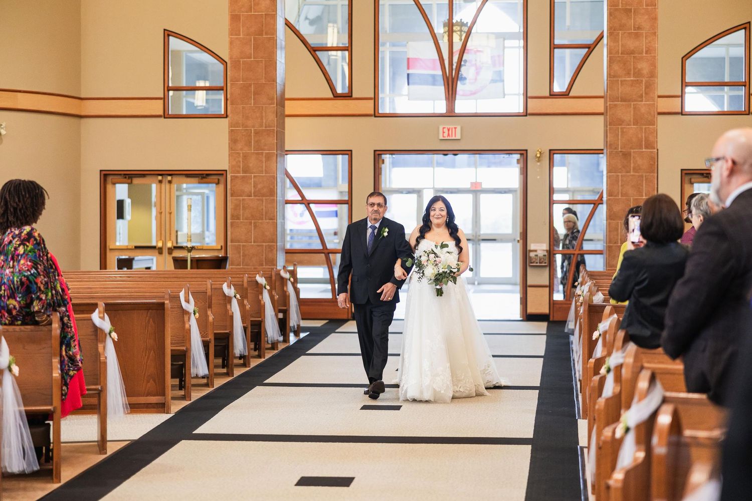 A wedding procession walking down the aisle of a modern church with high windows and wooden pews.