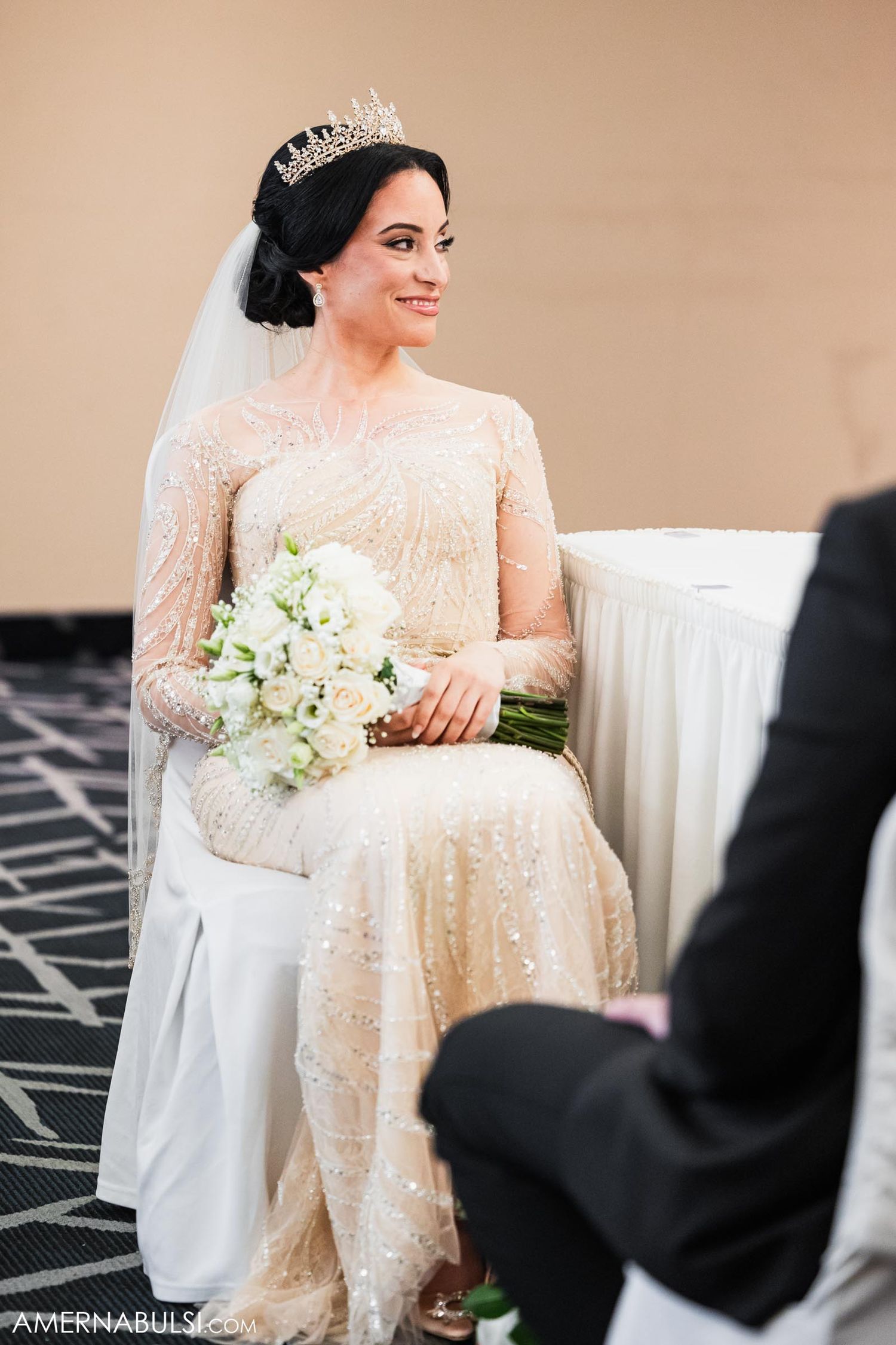 Bride in long-sleeved beaded gown holds white bouquet while seated during ceremony.