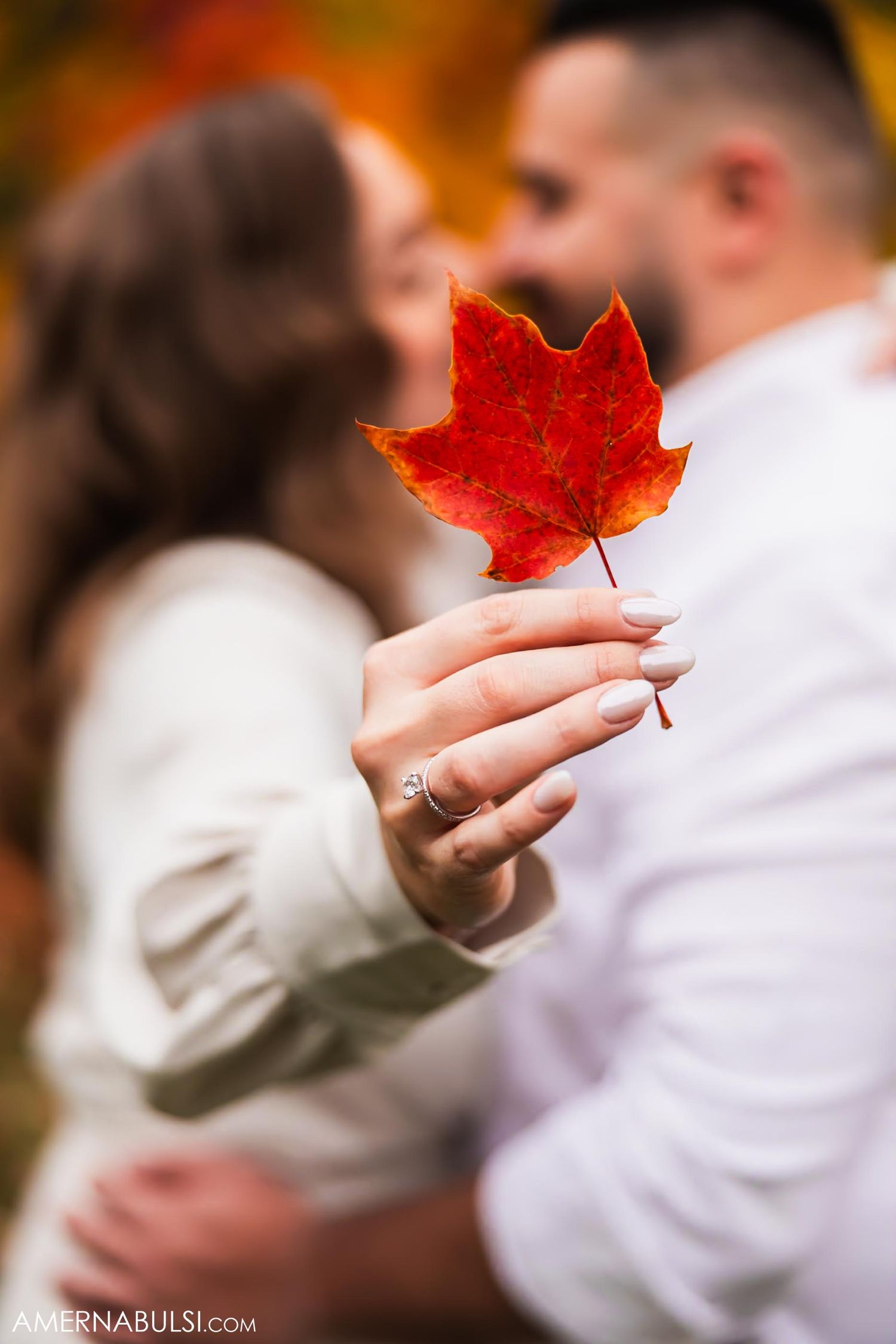 Close-up of hands holding a bright red maple leaf with an engagement ring visible.
