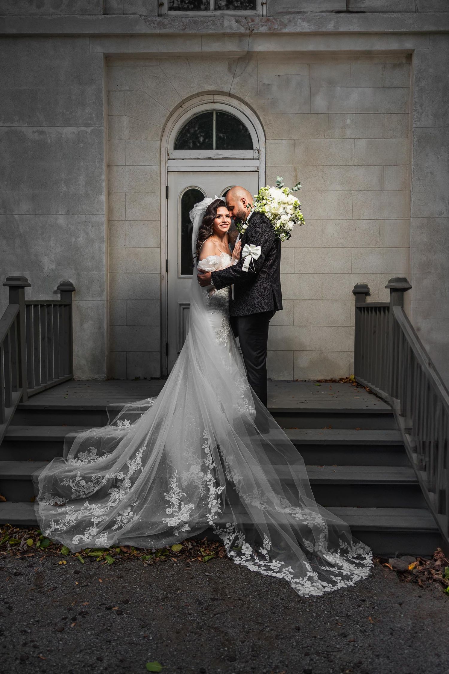 A couple embraces on stone steps in front of an elegant building with a stunning lace train.
