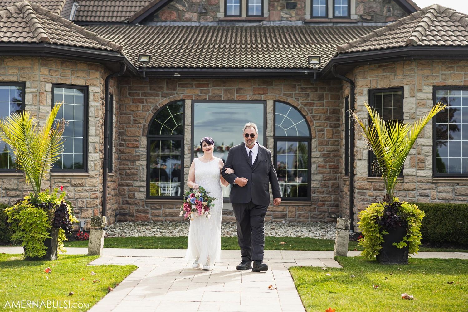 Wedding couple posing in front of stone mansion with landscaped entrance.