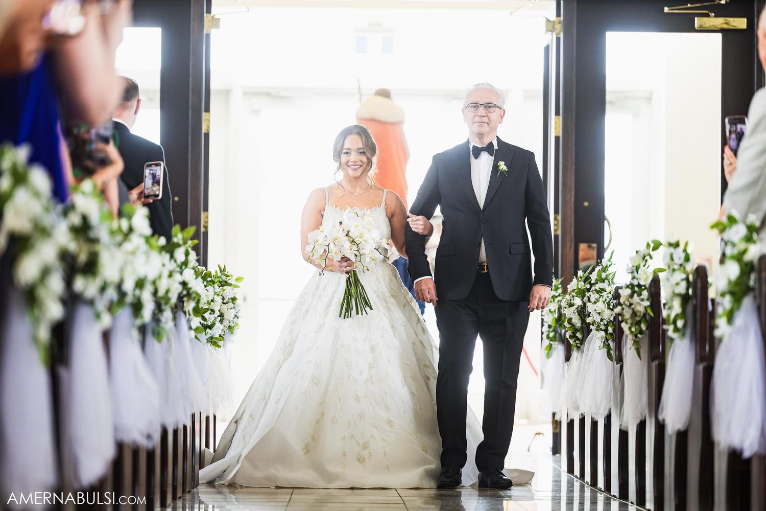 Bride and father walking down church aisle decorated with white floral arrangements.