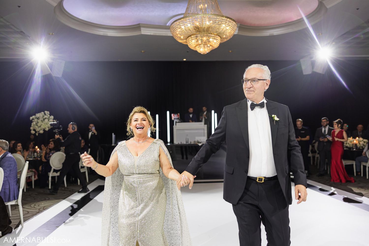 A couple in formal attire dances together under crystal chandeliers at an elegant wedding reception.