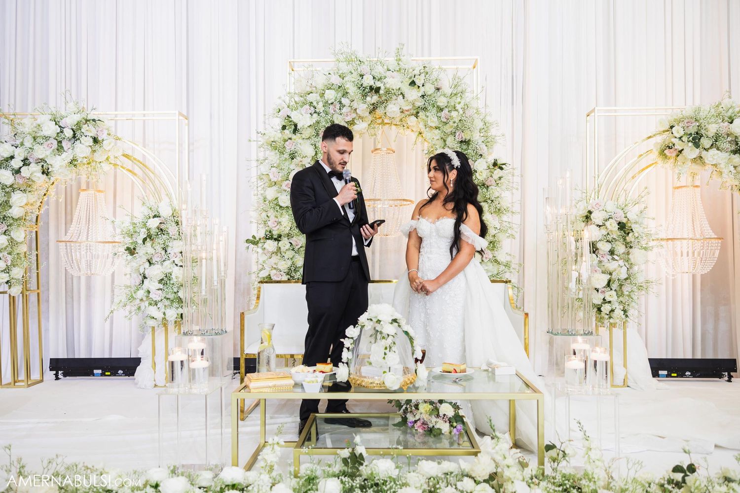 Wedding couple poses in front of an elegant white floral arch and gold decor.