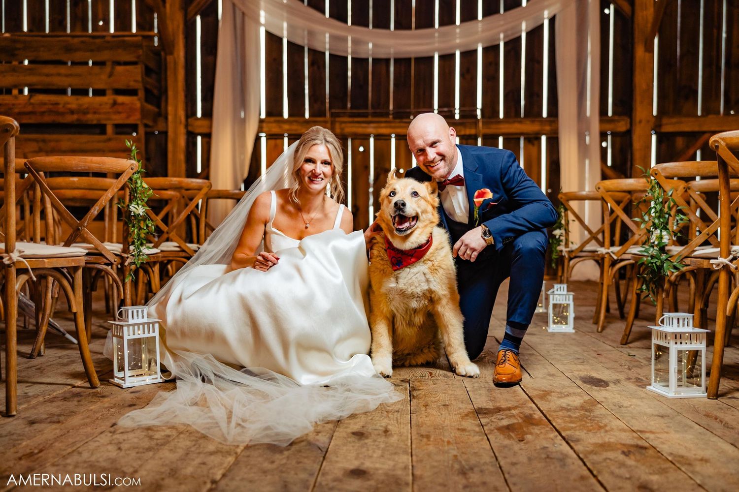 Couple poses with golden retriever in rustic barn setting surrounded by lantern decorations.