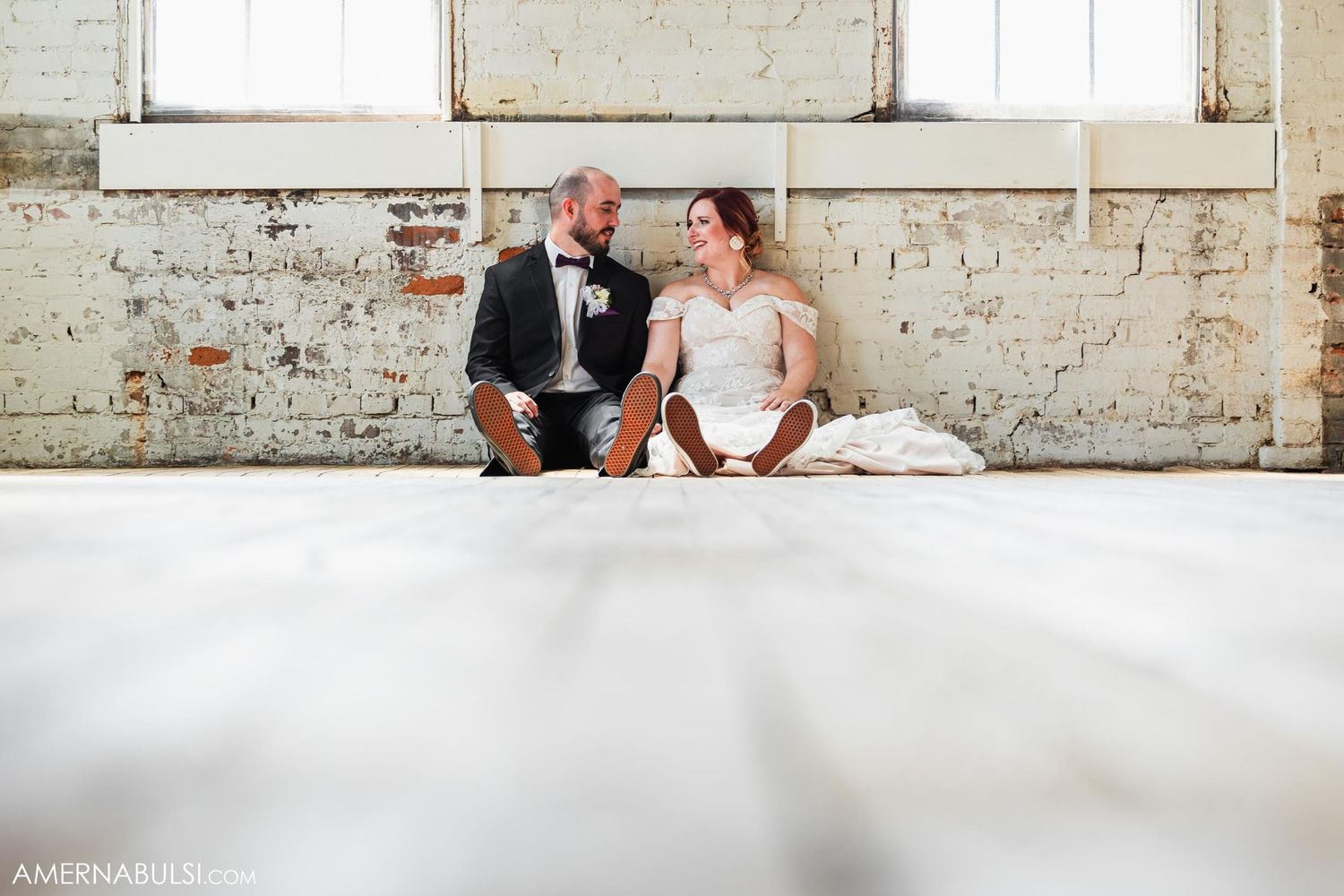 Wedding couple sits casually against white brick wall in industrial space.