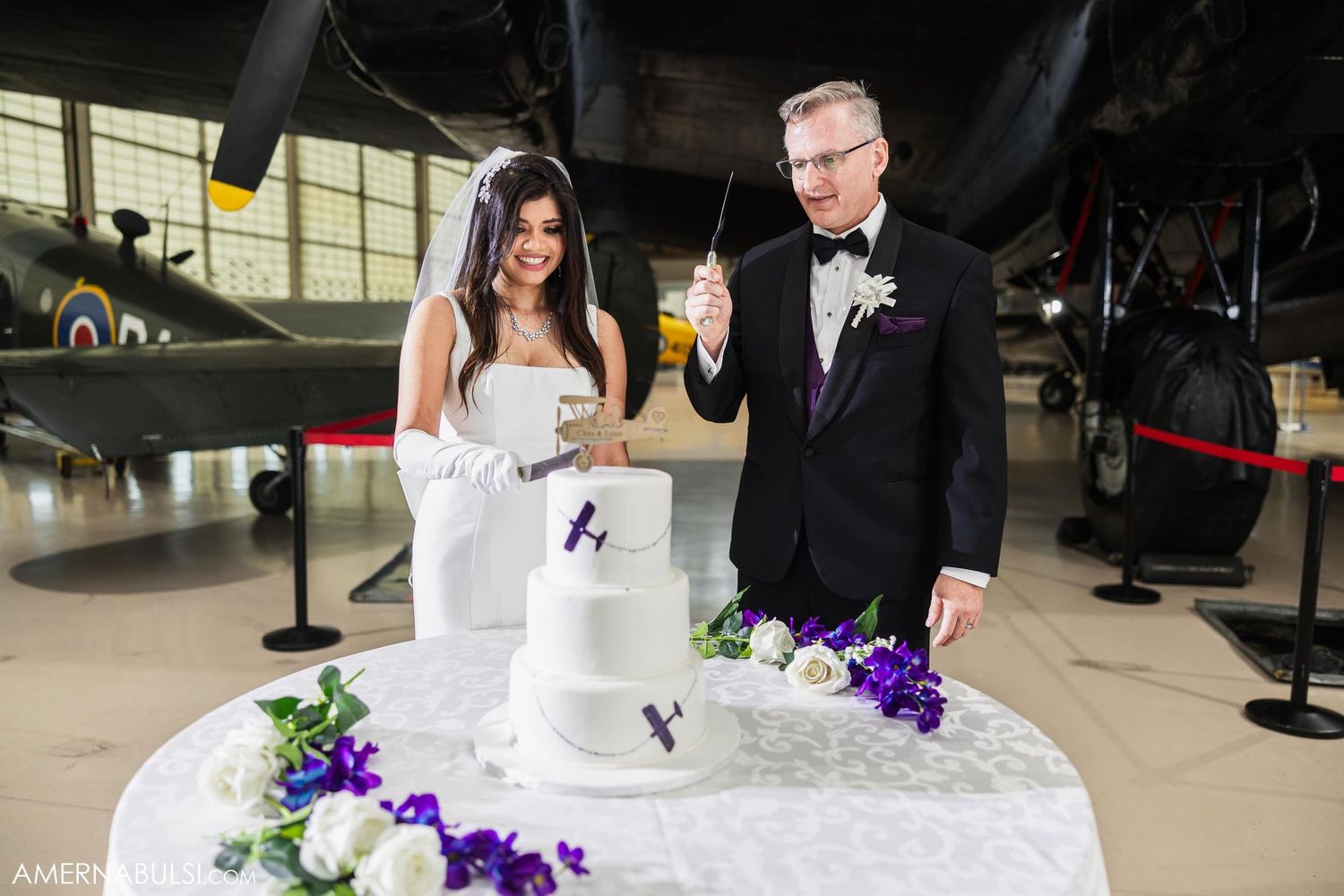 Newlyweds toast beside their aviation-themed white wedding cake in museum hangar.