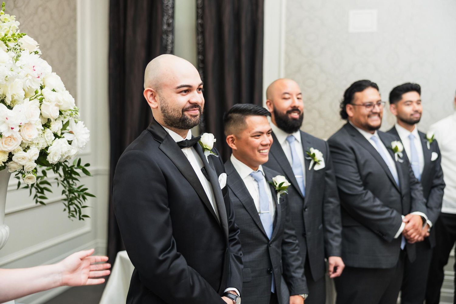 Groomsmen in formal attire stand together during wedding ceremony with white floral decor.