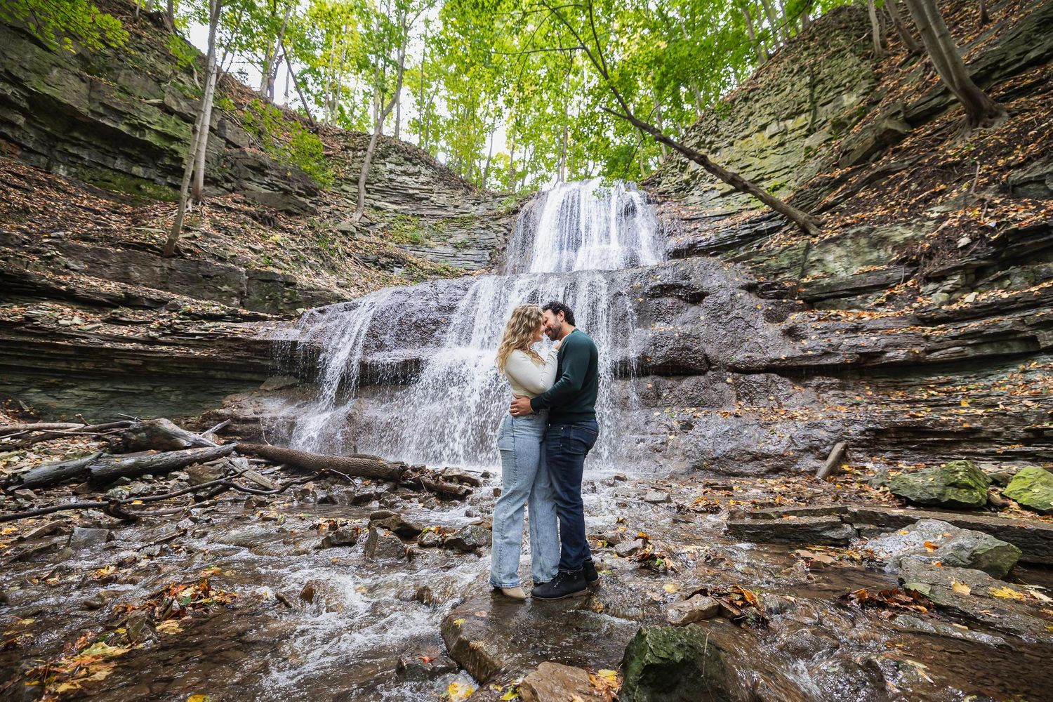 A couple embraces in front of a cascading waterfall surrounded by rocky cliffs.