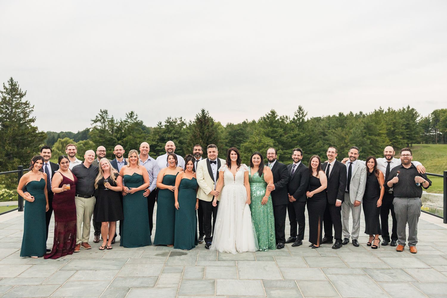 Wedding party in green and burgundy gowns with groom and groomsmen in black suits posing outdoors.